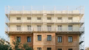 Edificio residencial español en obras de rehabilitación energética con andamios y aislamiento térmico de fachada en la mitad superior, ilustrativo del Plan Estatal de Vivienda 2026-2030 y el Plan Social para el Clima en comunidades de propietarios
